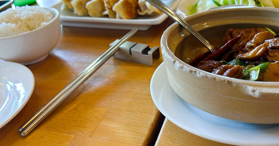 Stainless steel flatware rest holding a pair of metal chopsticks on a wooden restaurant table, beside a bowl of white rice and a clay pot dish with chicken and vegetables.