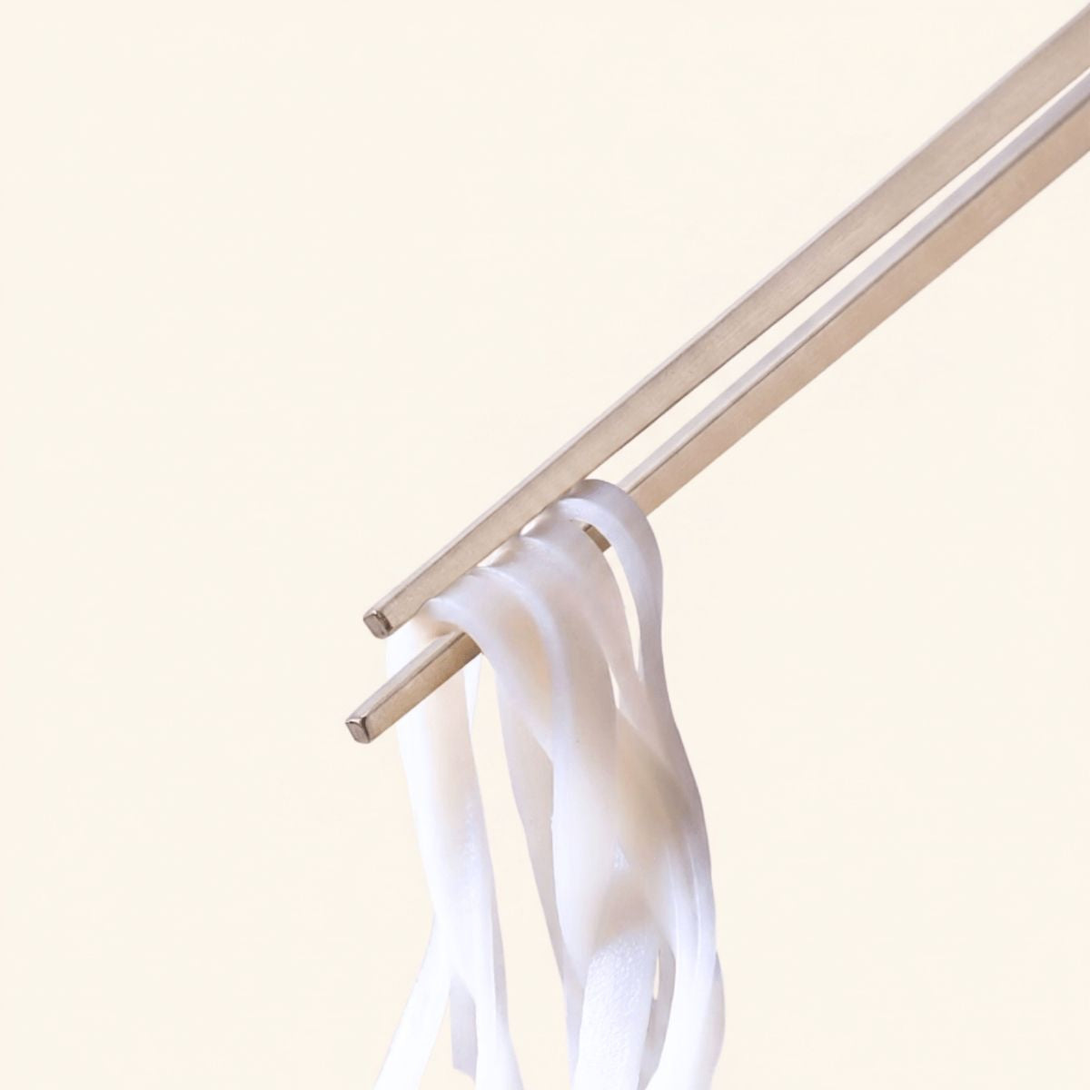 Radiko 316 stainless steel chopsticks holding flat white rice noodles, featuring square non-slip metal tips against a minimalist beige backdrop.