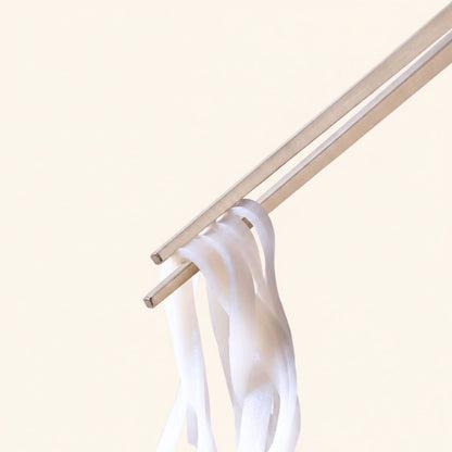 Radiko 316 stainless steel chopsticks holding flat white rice noodles, featuring square non-slip metal tips against a minimalist beige backdrop.