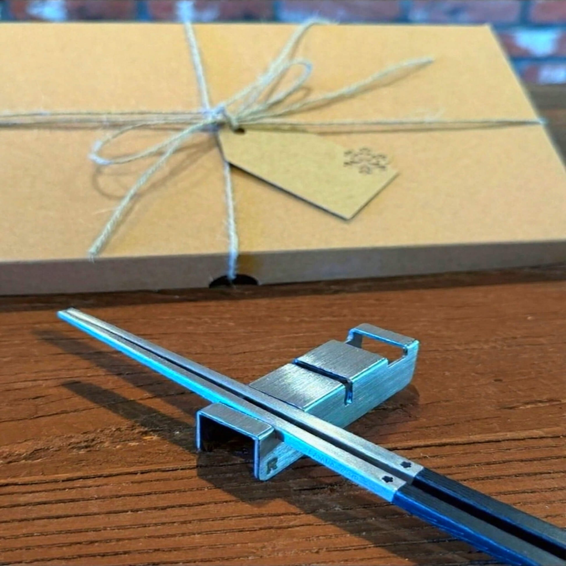 316 stainless steel chopsticks with 304 utensil rest set in the front with Radiko 100% recyclable brown kraft packaging box in the back on a brown wood table
