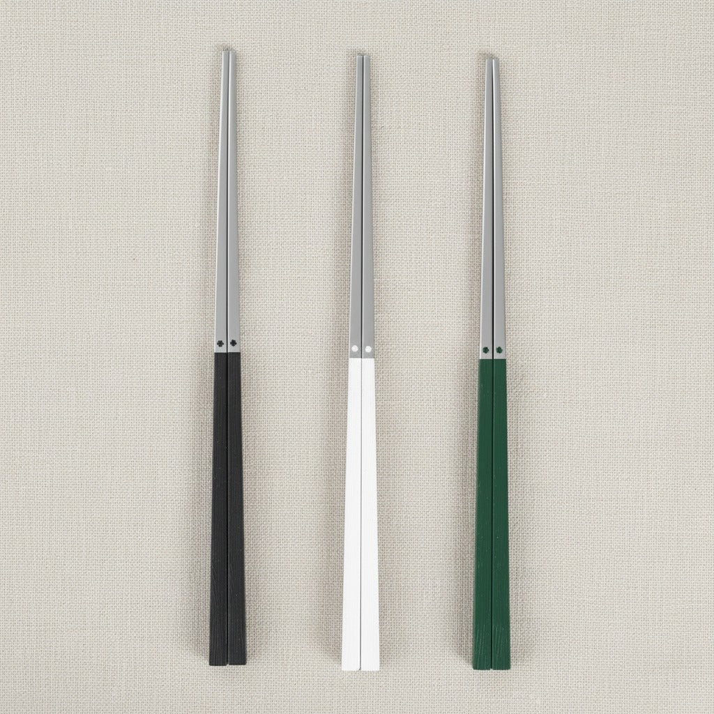 Three pairs of chopsticks with silver metal tips and handles in black, white, and green, arranged vertically on a textured beige fabric background.