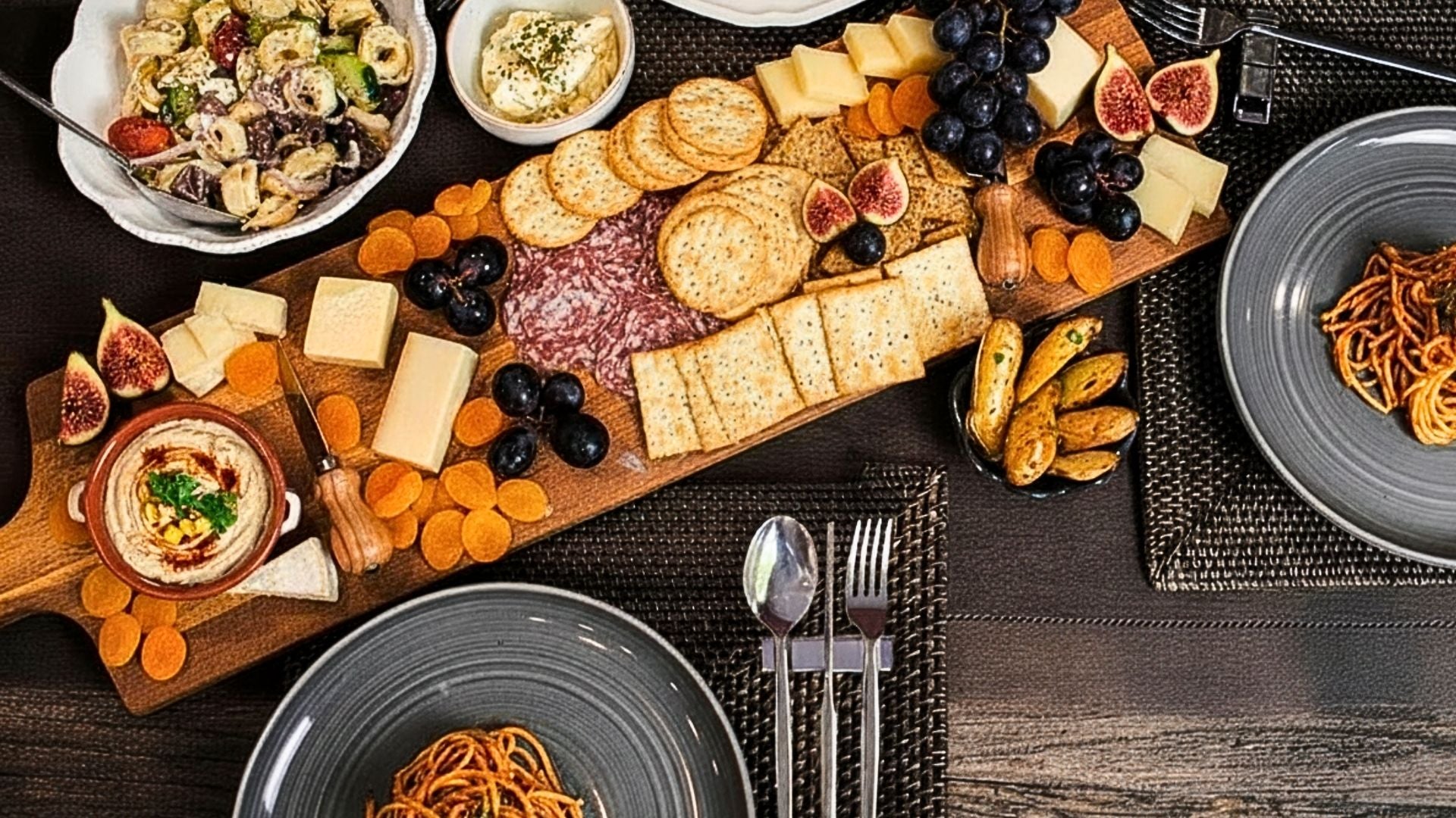 Grazing board with cheese, crackers, grapes, figs, and hummus on a wooden platter, styled on a dark table with Radiko 304 stainless steel flatware rest.